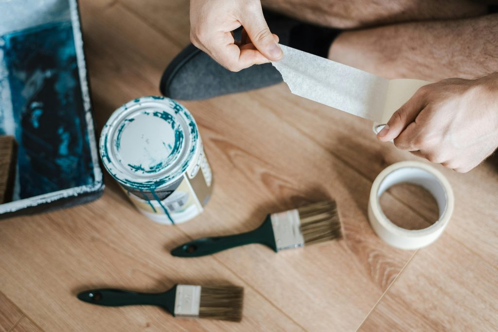 Close up of person on the floor with teal paint, two paint brushes, and painter's tape.