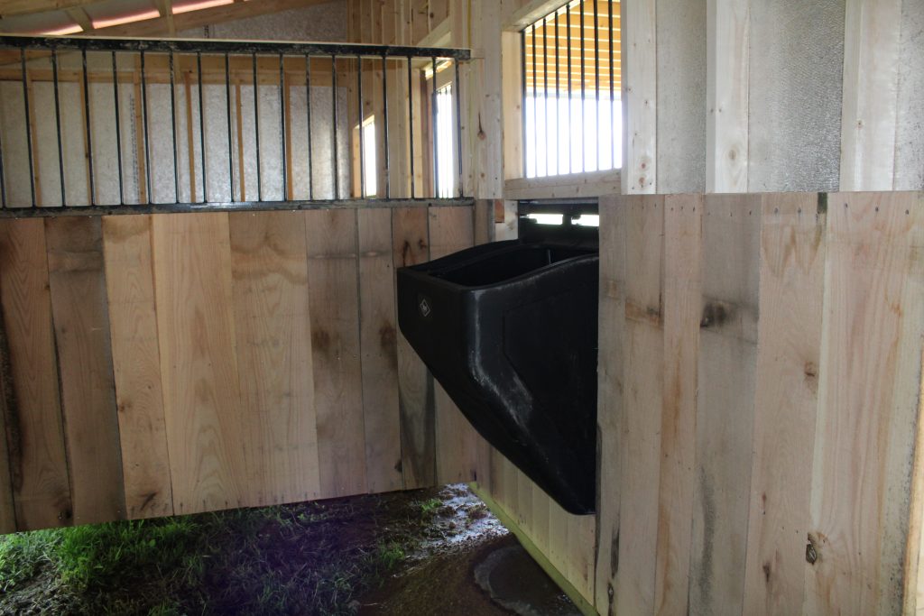 Interior of a monitor horse barn stall with wood kickboards, no floor, and a black horse feeder.