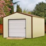Storage building with beige siding, brown trim, a brown metal roof, and a white roll-up garage door with a ramp sitting in a fenced-in backyard.