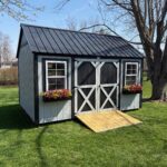 Pocahontas Shed with light gray siding, black trim, 2 windows and flower boxes, black double doors with a ramp, and a black metal roof.