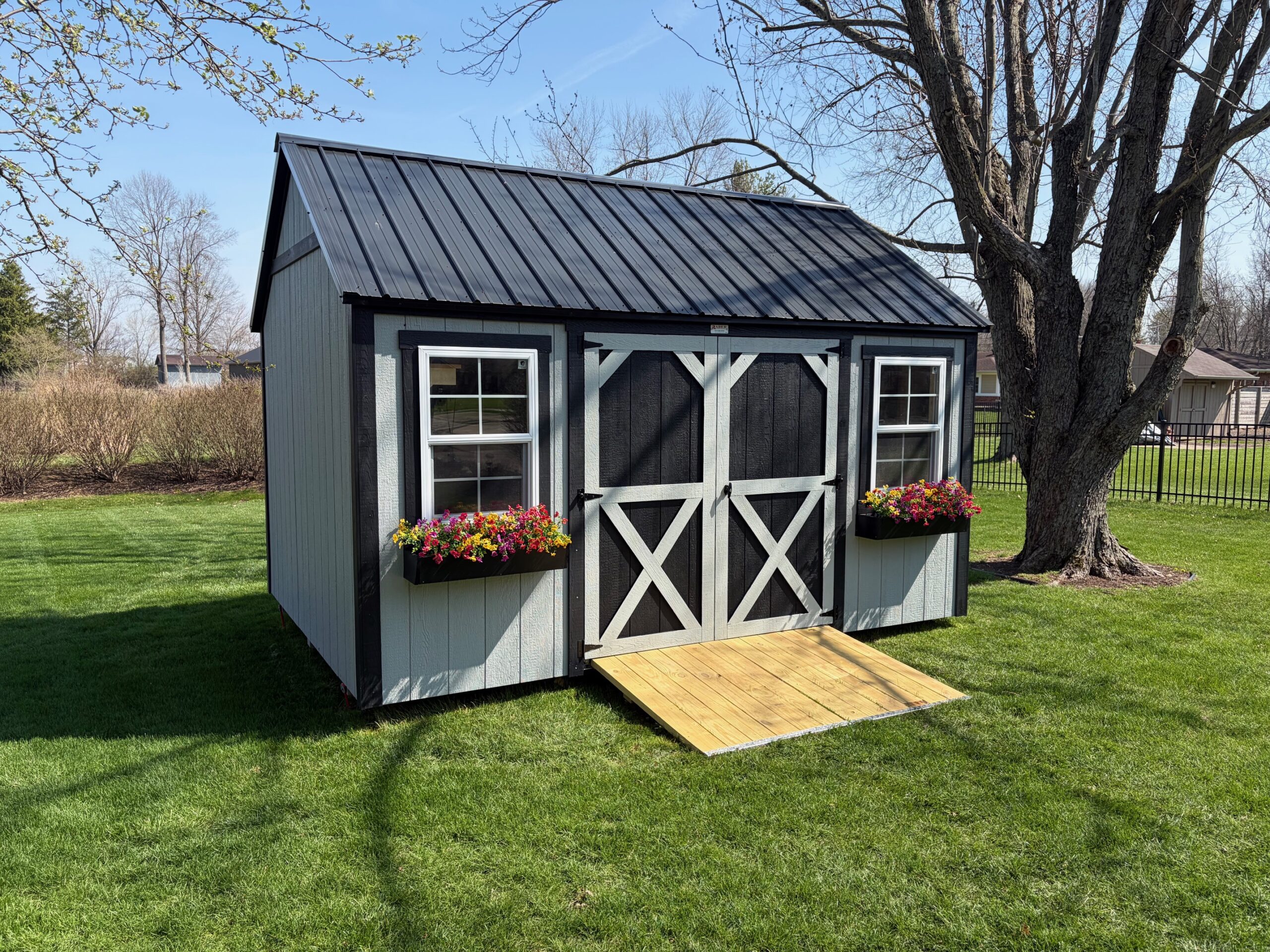 Pocahontas Shed with light gray siding, black trim, 2 windows and flower boxes, black double doors with a ramp, and a black metal roof.